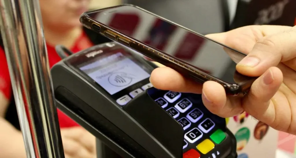 A customer holds a smartphone over a contactless payment terminal to complete a tap-to-pay transaction at a retail checkout, highlighting the convenience of digital and cashless payments in a modern business environment.
